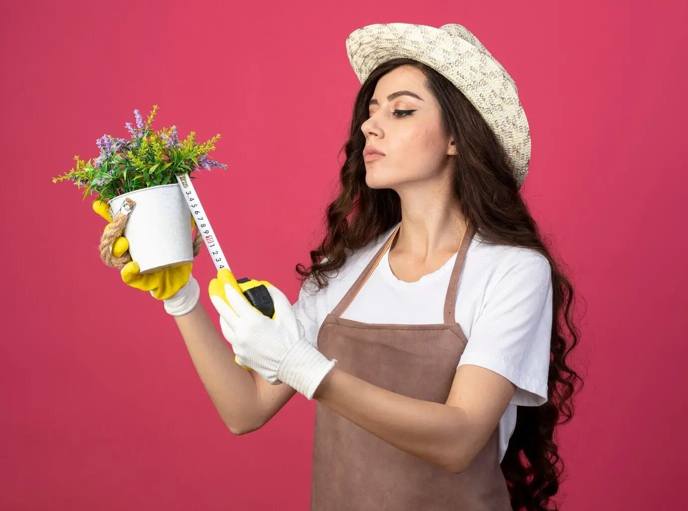 A confident young female gardener in uniform, wearing a gardening hat, measures a flowerpot with a tape measure, isolated against a pink wall with copy space.