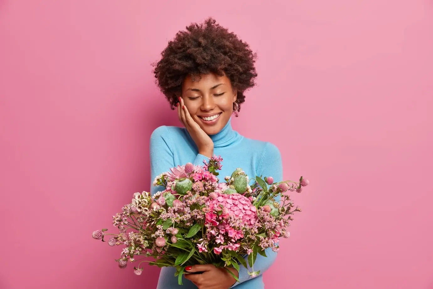 Photo of a pleased, touched young Afro-American woman who received an anniversary gift, holding a large bouquet of flowers, eyes closed and smiling gently, wearing a blue turtleneck.
